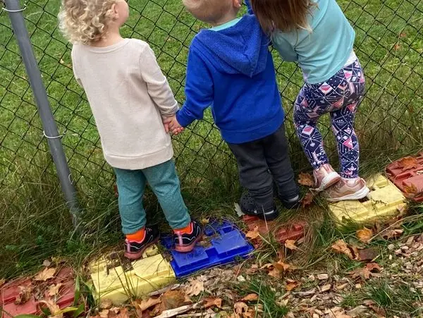 Three young children stand holding hands on colorful plastic stepping blocks beside a chain-link fence in a grassy outdoor area with fallen leaves.