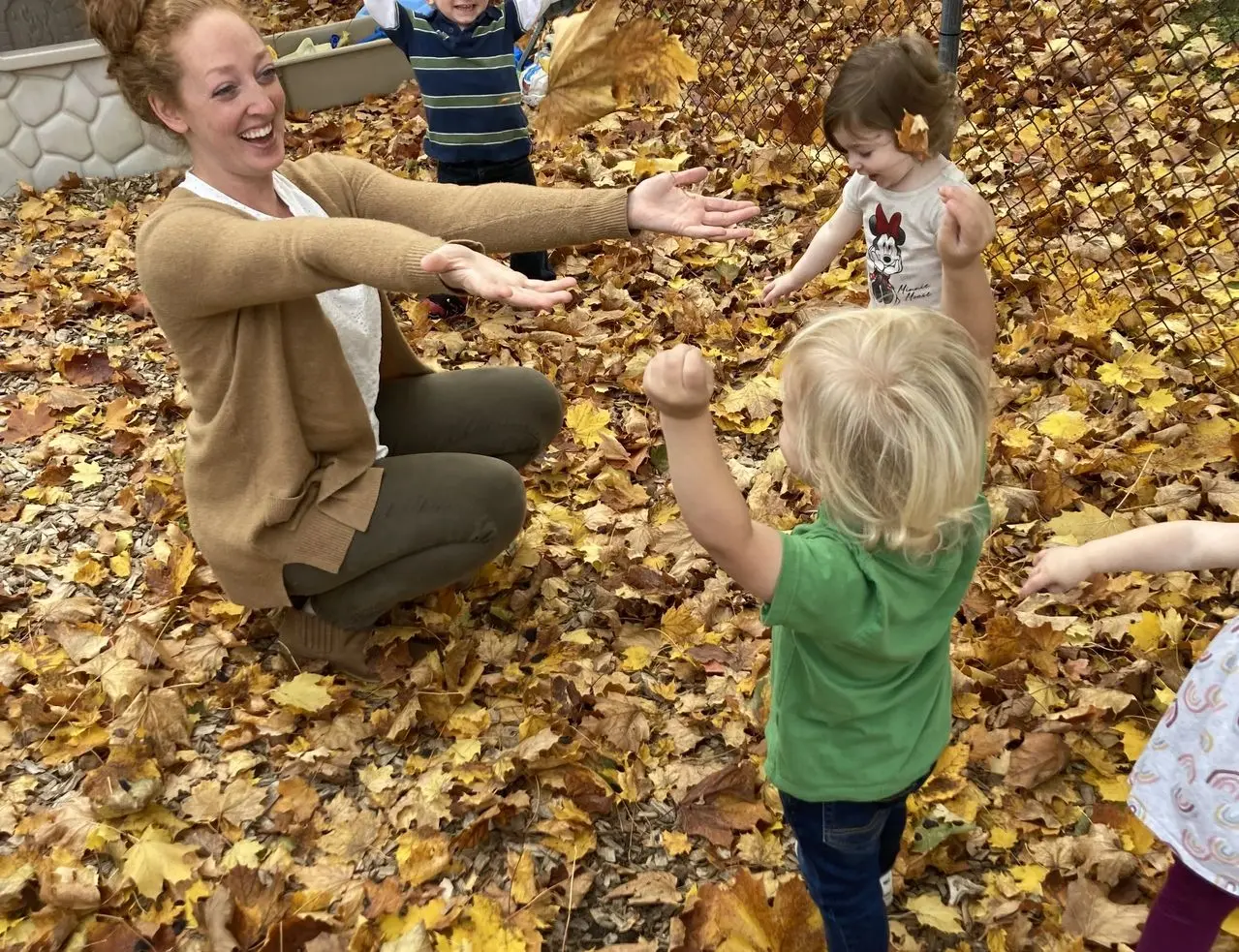 A woman crouches among fallen leaves with her arms outstretched while several young children stand around her, one tossing leaves into the air.
