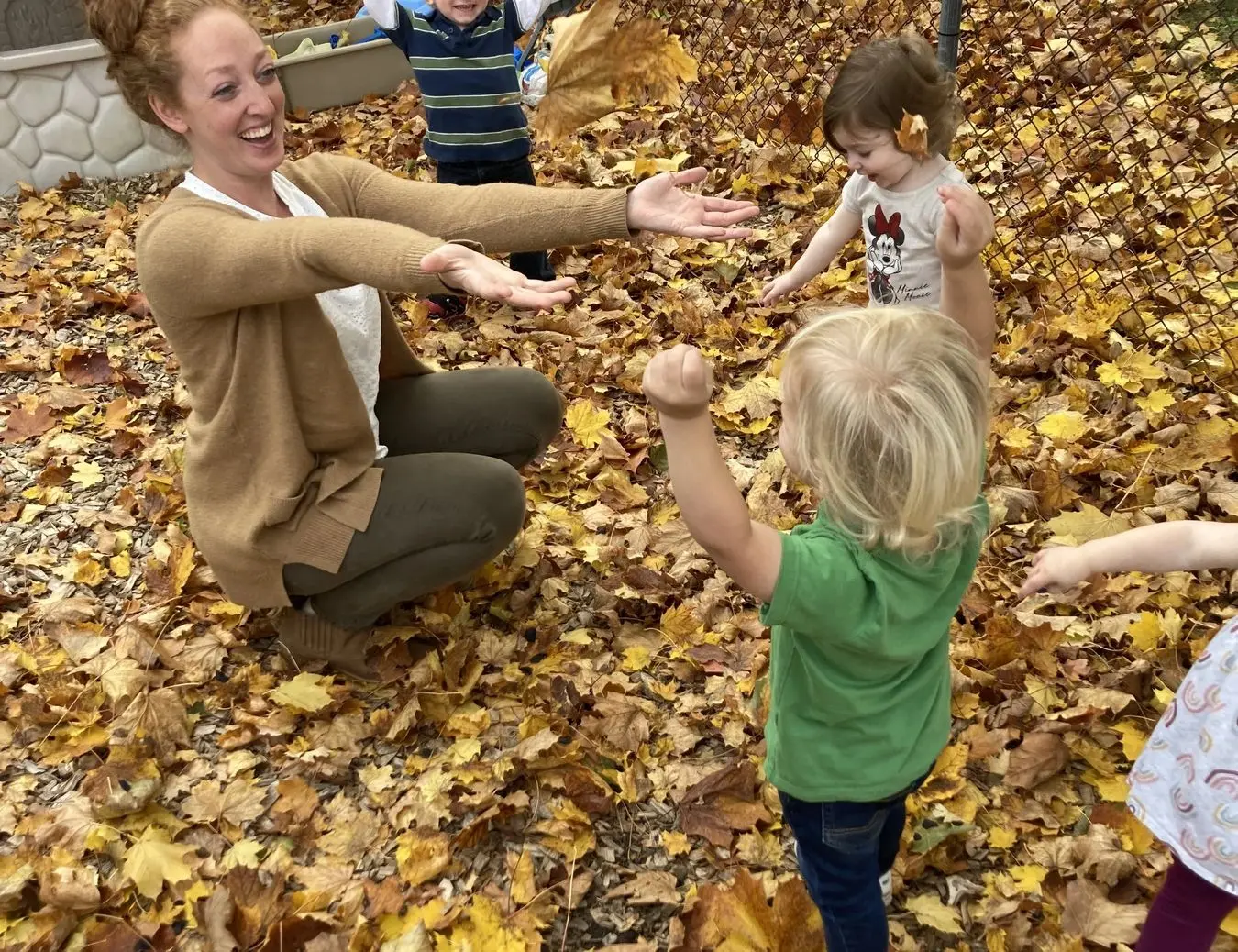 A woman crouches among fallen leaves with her arms outstretched while several young children stand around her, one tossing leaves into the air.
