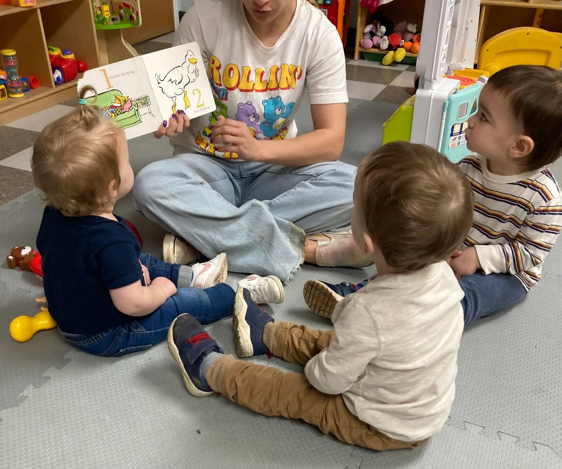 An adult sits cross-legged on a mat holding an open picture book while three young children sit facing them in a play area with toy shelves.