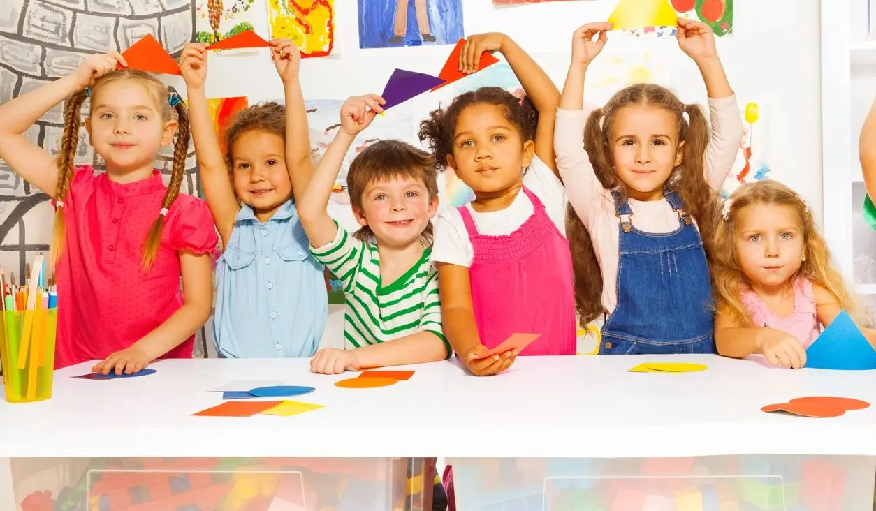 Six children stand behind a white table holding colored paper shapes, with crayons and paper pieces on the table and artwork on the wall behind them.