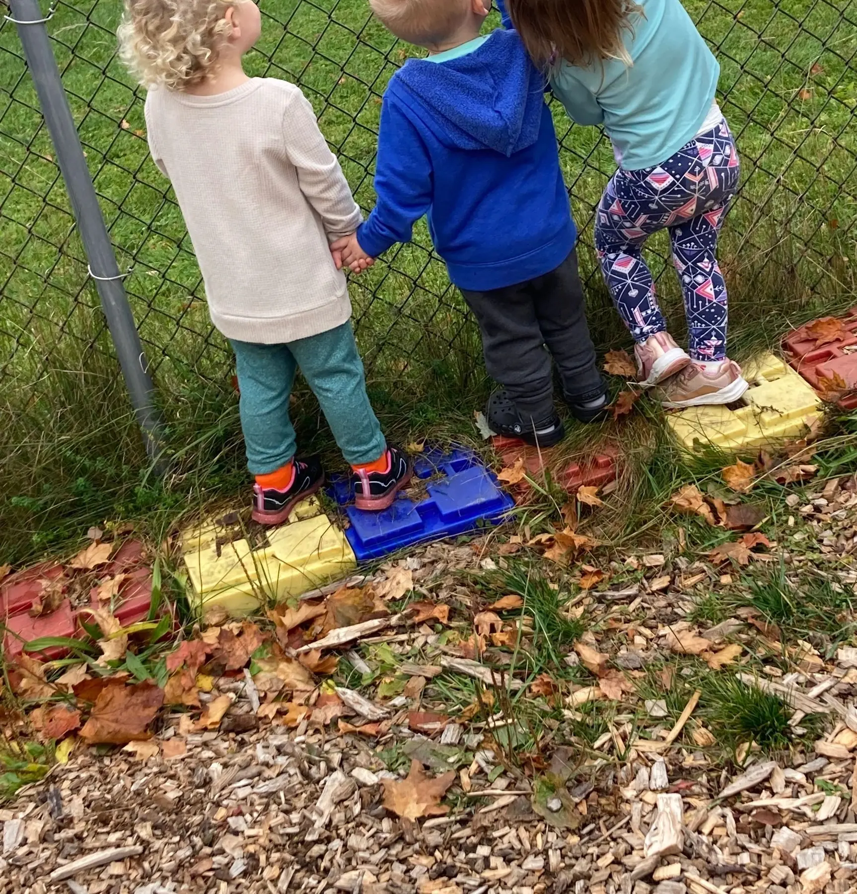 Three young children stand on colorful plastic blocks beside a chain-link fence, with two holding hands while the third climbs, surrounded by fallen leaves and wood chips.