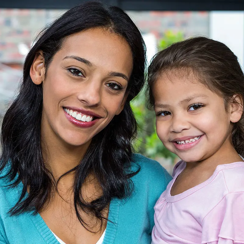 A woman in a teal cardigan and a young girl in a pink top smile side by side facing the camera.