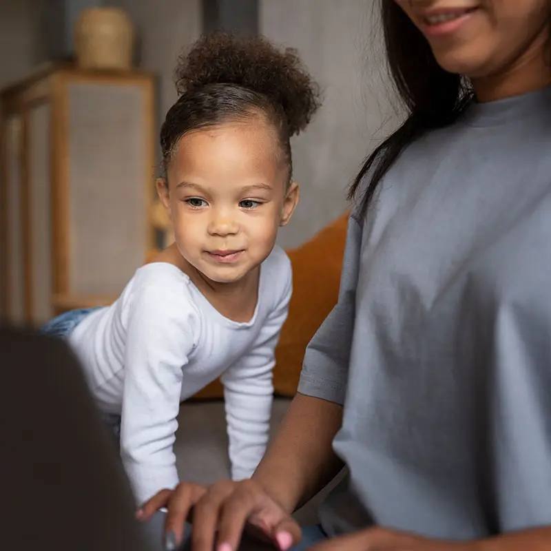 A toddler in a white long-sleeve shirt leans forward on hands while a seated person in a gray shirt sits beside them indoors.