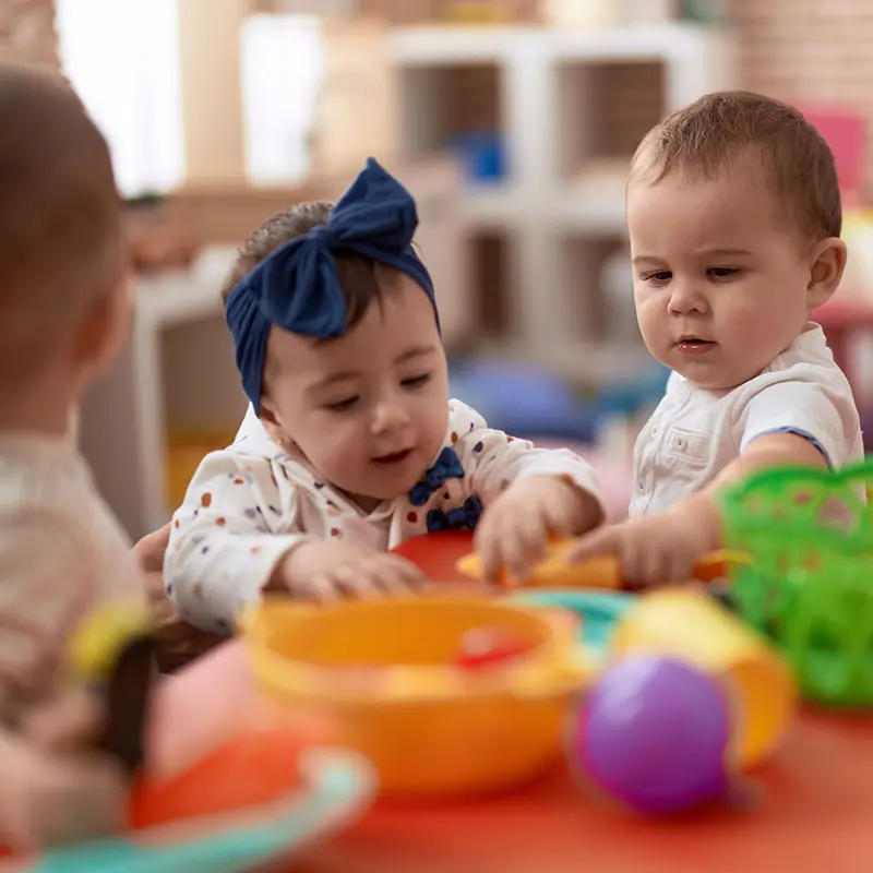 Three babies sit at a table with colorful toys, including a yellow bowl and a purple ball, while one baby in a blue bow headband reaches toward the toys.