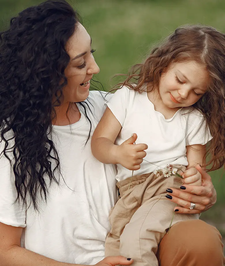A woman with long dark curly hair smiles while holding a young girl with wavy brown hair who is looking down at a small sprig of white flowers in her hands.