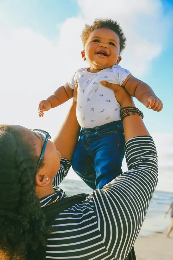 An adult with braided hair and glasses holds a smiling baby in a white patterned shirt and blue jeans up in the air against a blue sky.