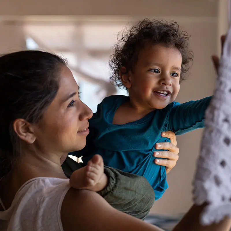 A woman holds a smiling toddler who reaches toward a white crocheted object on the right side of the frame.