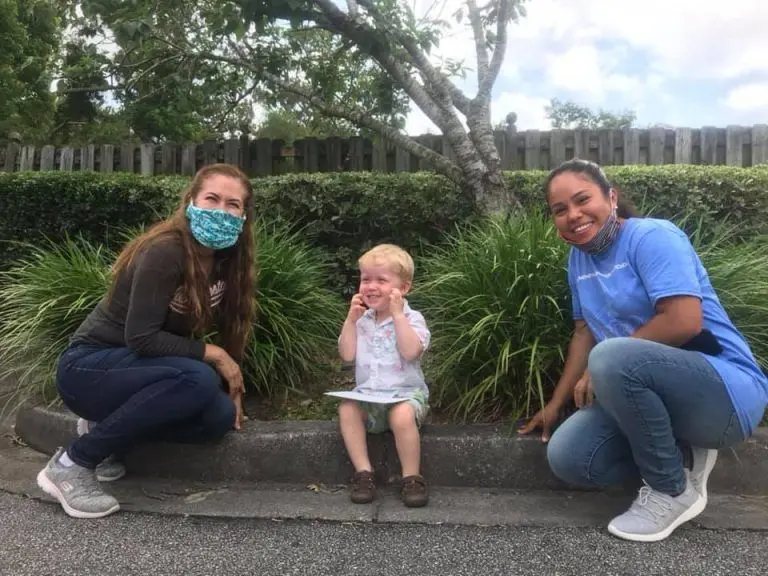 Two women wearing face masks crouch on either side of a young boy sitting on a concrete curb, holding a piece of paper and touching his face.