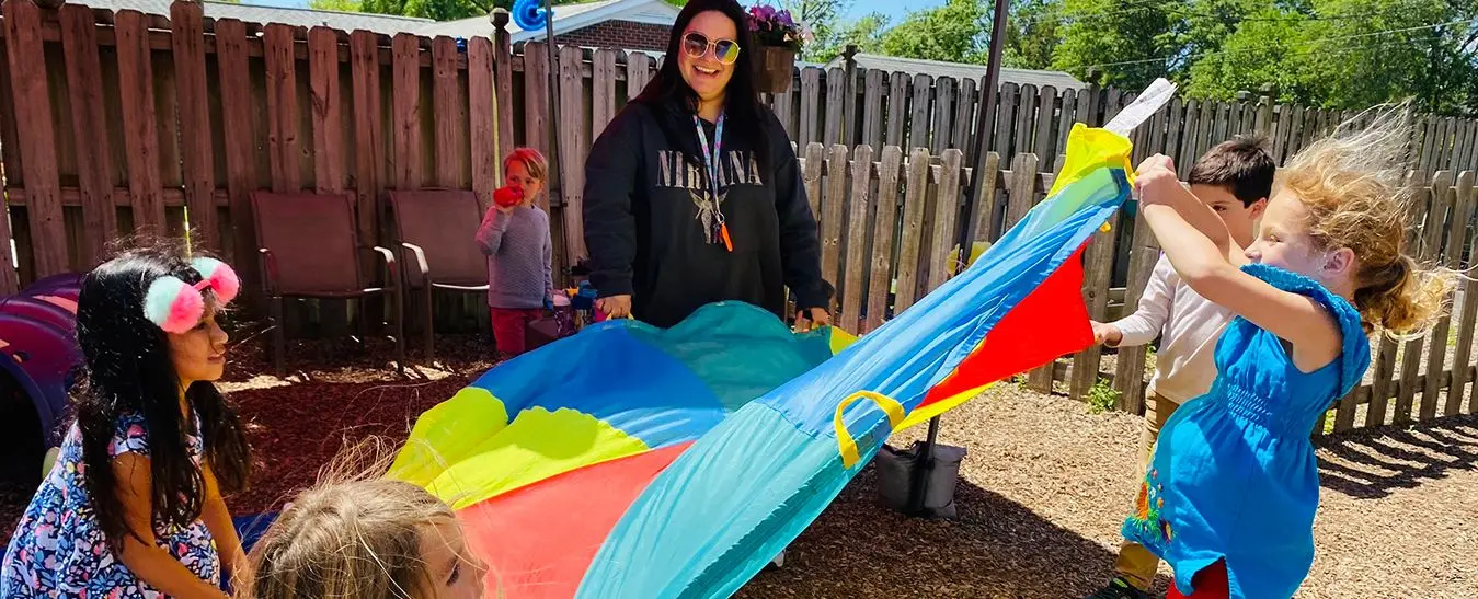 Several children and an adult stand on a playground holding the edges of a large multicolored parachute.