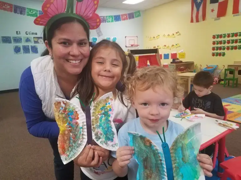A woman and two children hold painted paper butterfly crafts while another child paints at a table in a classroom decorated with flags and wall displays.