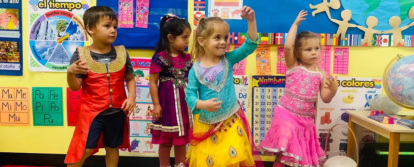 Four children in colorful costumes stand in front of a classroom wall decorated with educational posters and a globe.