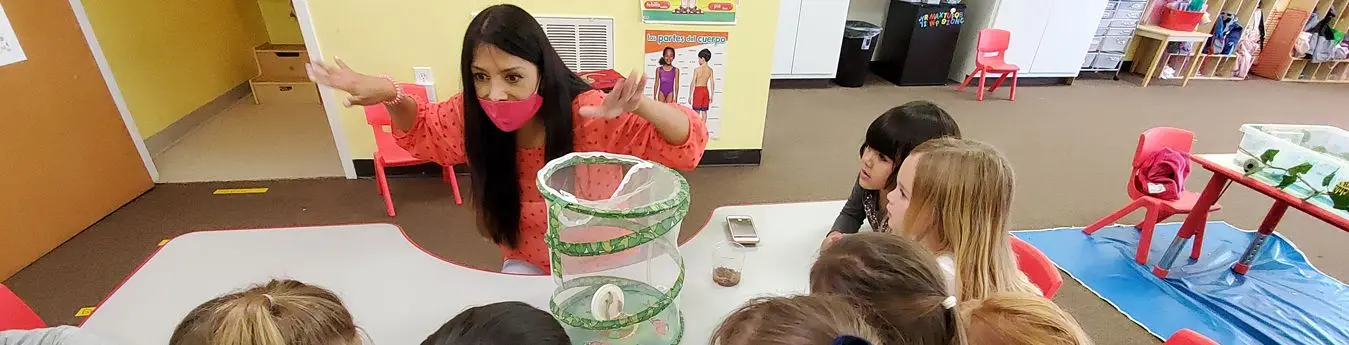 A woman in a pink mask gestures with both hands beside a mesh insect habitat on a table while several children watch.