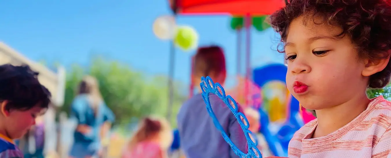 A young child with curly hair purses their lips while holding a blue beaded craft item, with other children and colorful objects blurred in the background.