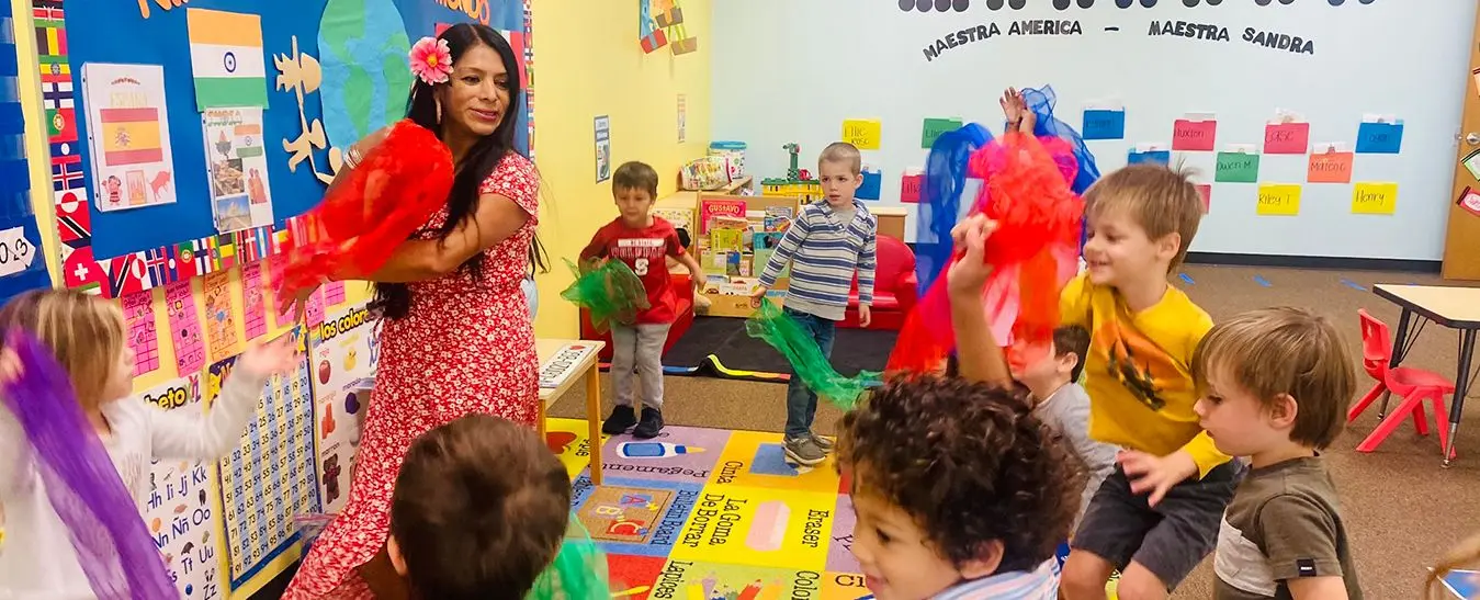 A teacher in a red floral dress holds red scarves while several young children dance and play with colorful scarves on a patterned classroom rug.