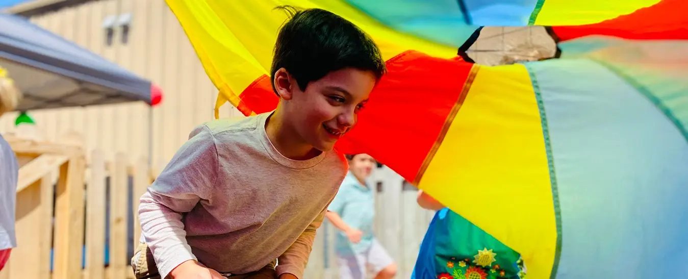 A smiling boy in a gray shirt leans forward while holding the edge of a large multicolored parachute as other children play behind it.