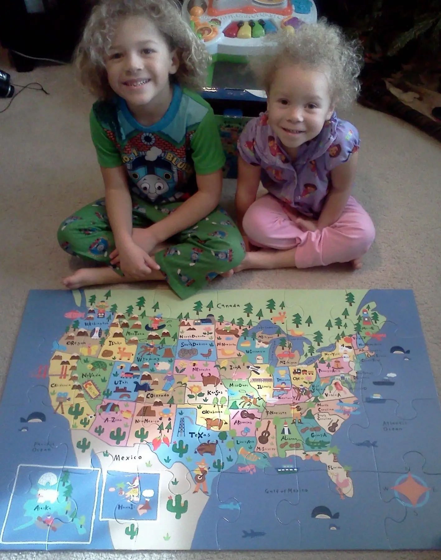 Two children sit on a carpeted floor behind a completed United States map puzzle.