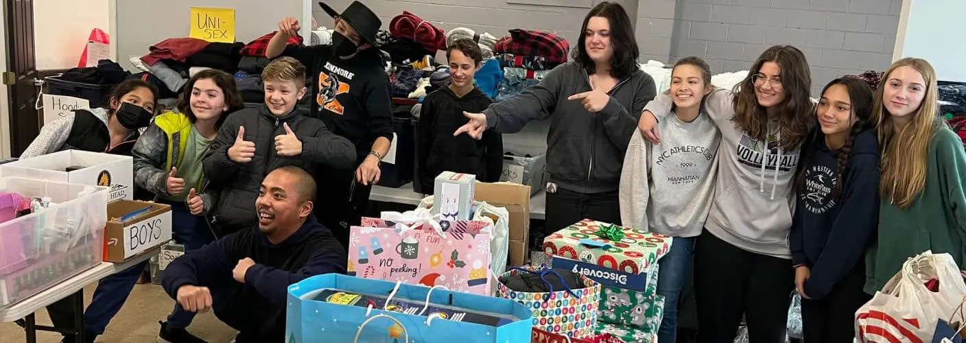 A group of children and adults pose together indoors among tables holding boxes and wrapped gifts, with several individuals pointing toward the camera.
