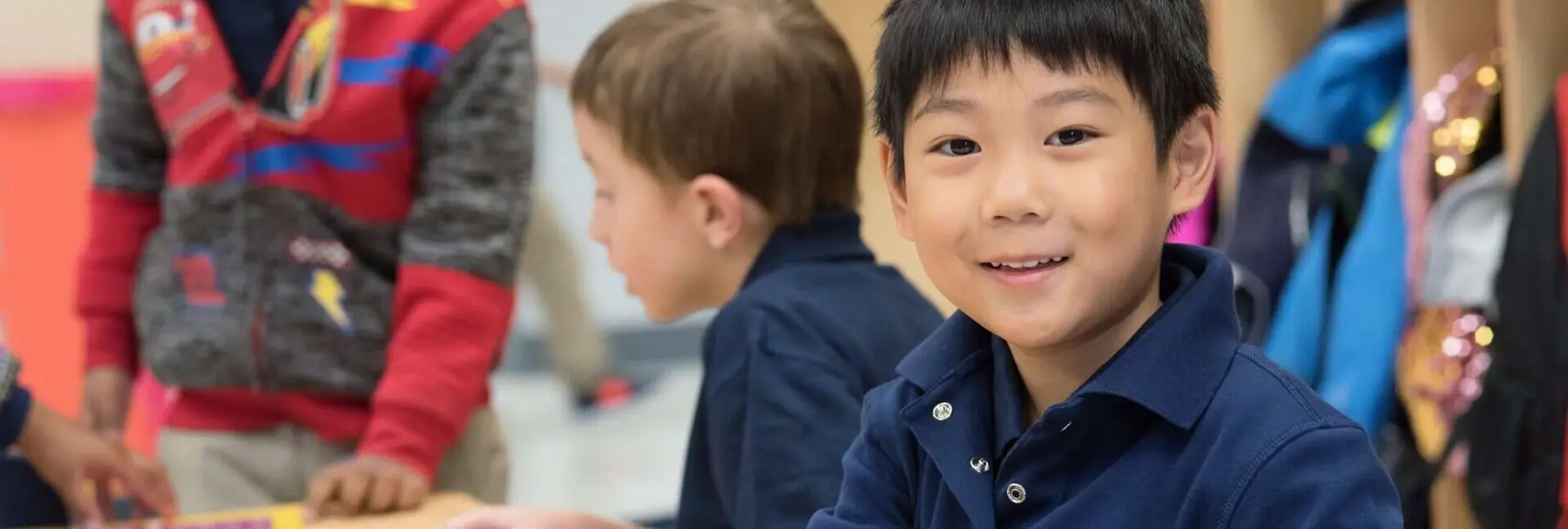 A boy in a navy polo shirt smiles at the camera while two other children stand near a table in the background.