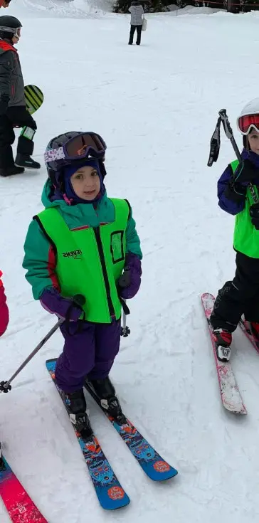 A child in a green vest and purple snow pants stands on skis holding poles on a snowy slope while another child in similar gear stands nearby.