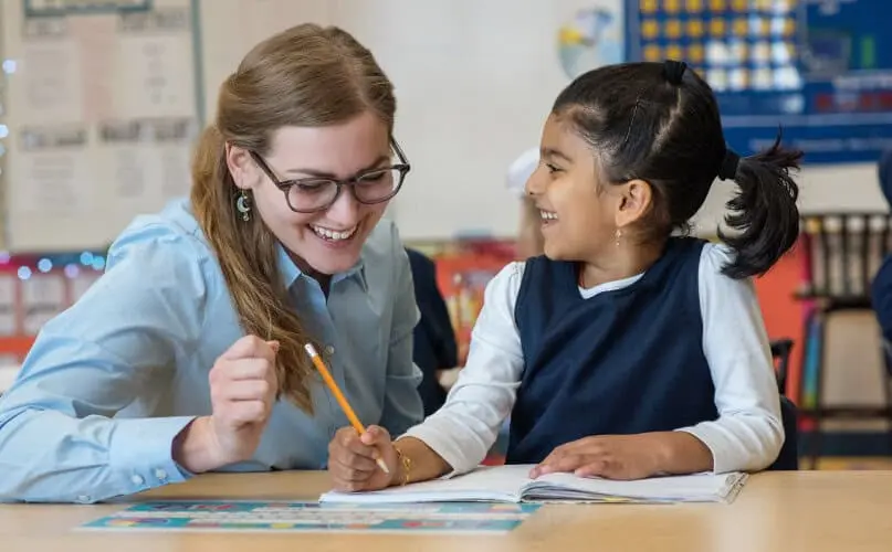 An adult woman with glasses and a girl in a navy vest sit at a table, smiling at each other while holding a pencil over an open notebook.