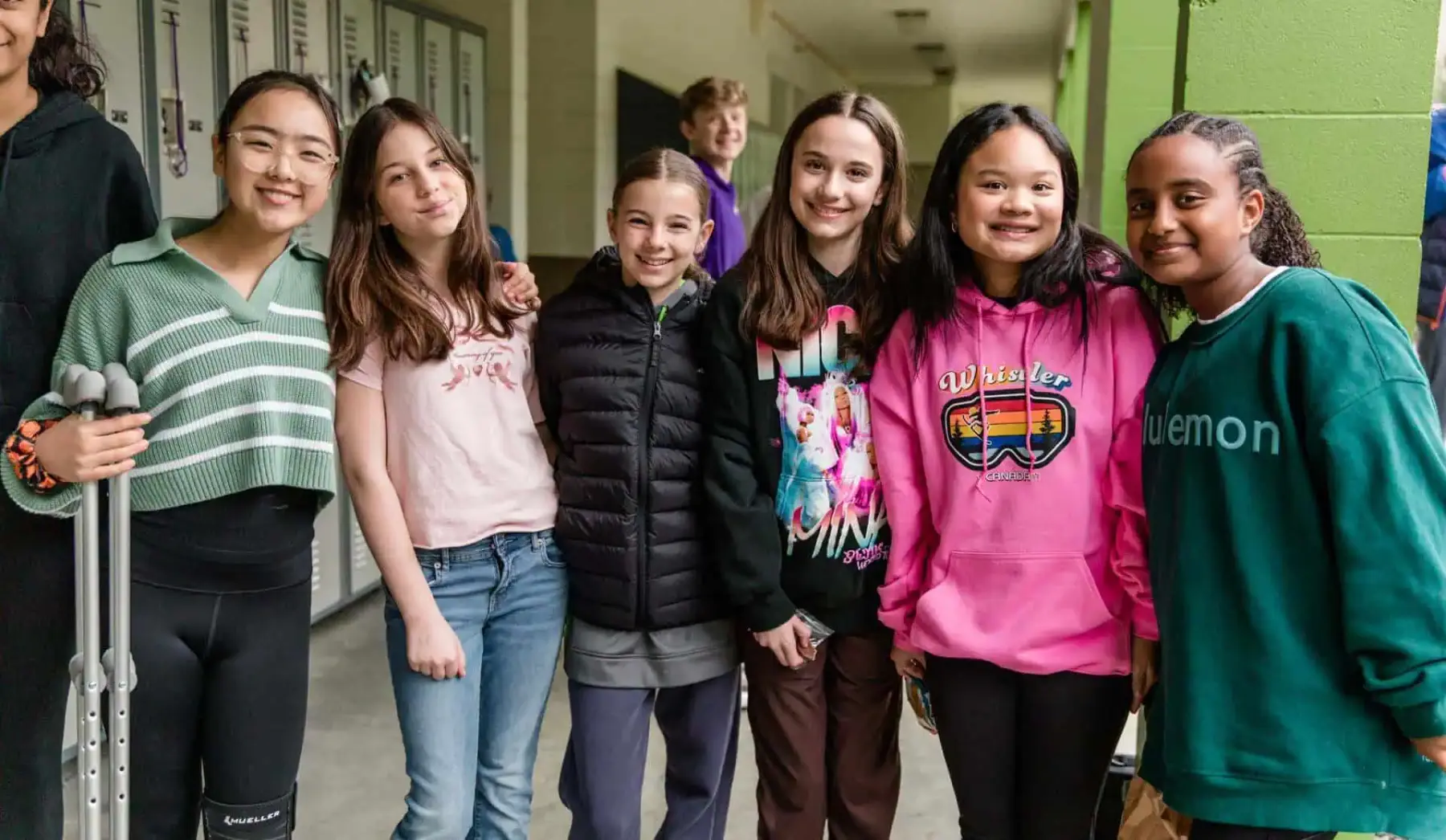 A group of seven children stand together in a hallway, one holding crutches, with lockers and a green wall visible behind them.