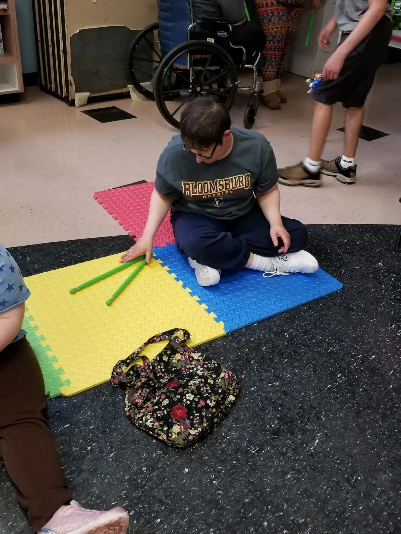 A boy in a dark t-shirt sits cross-legged on colorful foam mats holding two green sticks while other people stand nearby in a room with a wheelchair.