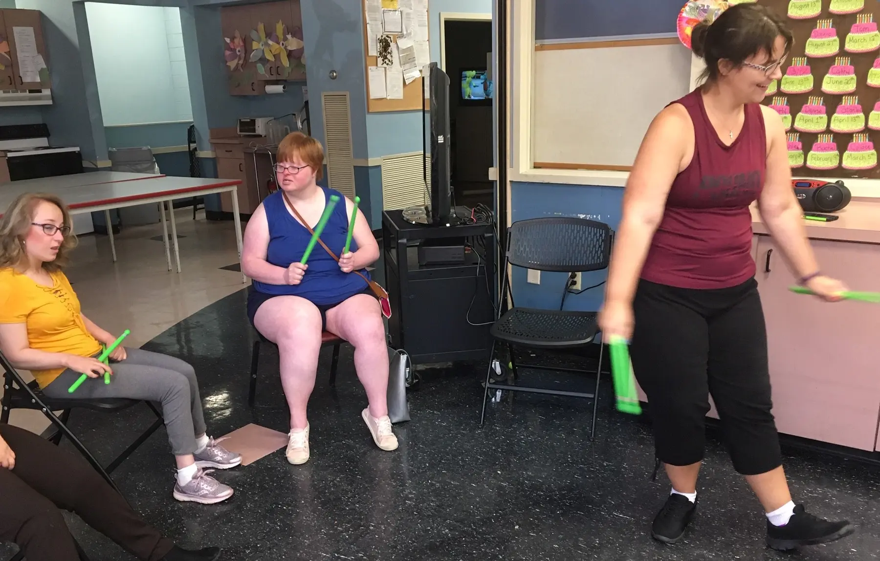 Three women sit and stand in a room holding green rhythm sticks, with a bulletin board of cupcake cutouts and a television on a cart in the background.