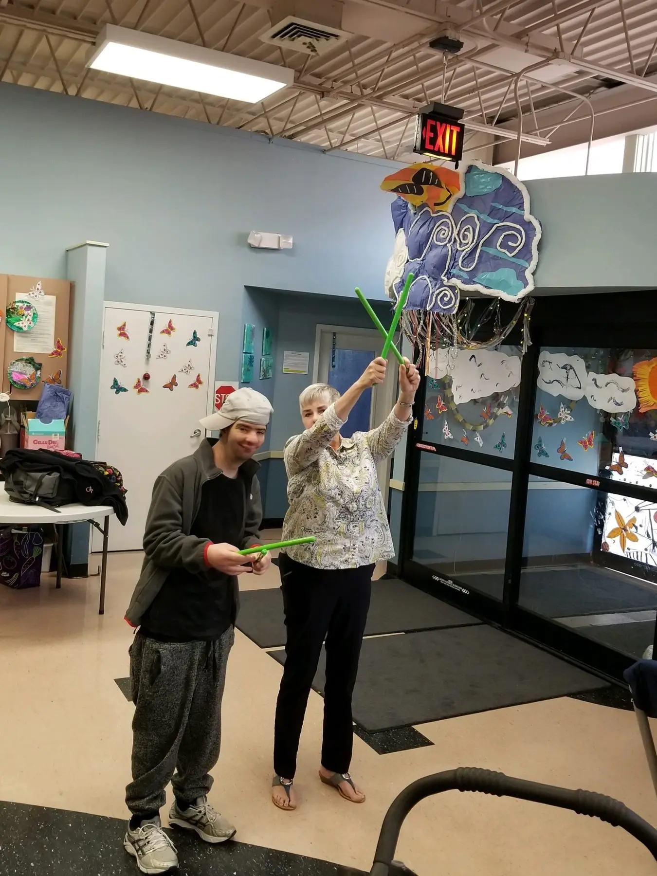 A young person in a cap and a woman stand indoors holding green sticks up toward a hanging paper craft near a glass door.