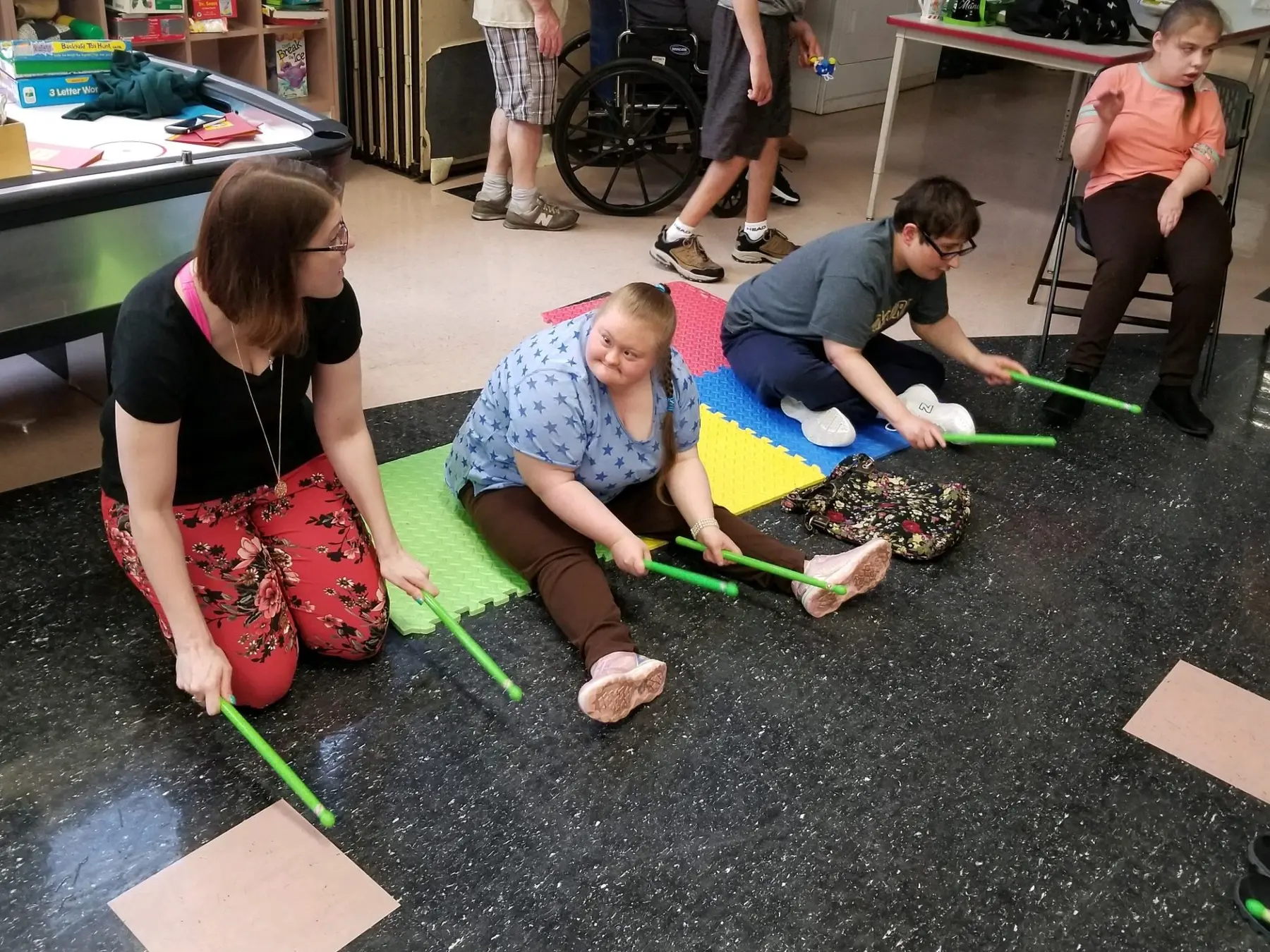 Four people sit or kneel on a dark speckled floor holding green rhythm sticks above paper squares and foam mats while a fifth person stands near a wheelchair in the background.