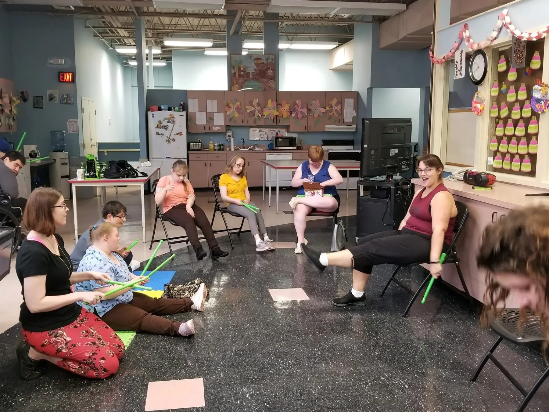 Several adults sit in a circle on chairs and the floor holding green rhythm sticks in a room with cabinets, a refrigerator, and a bulletin board.