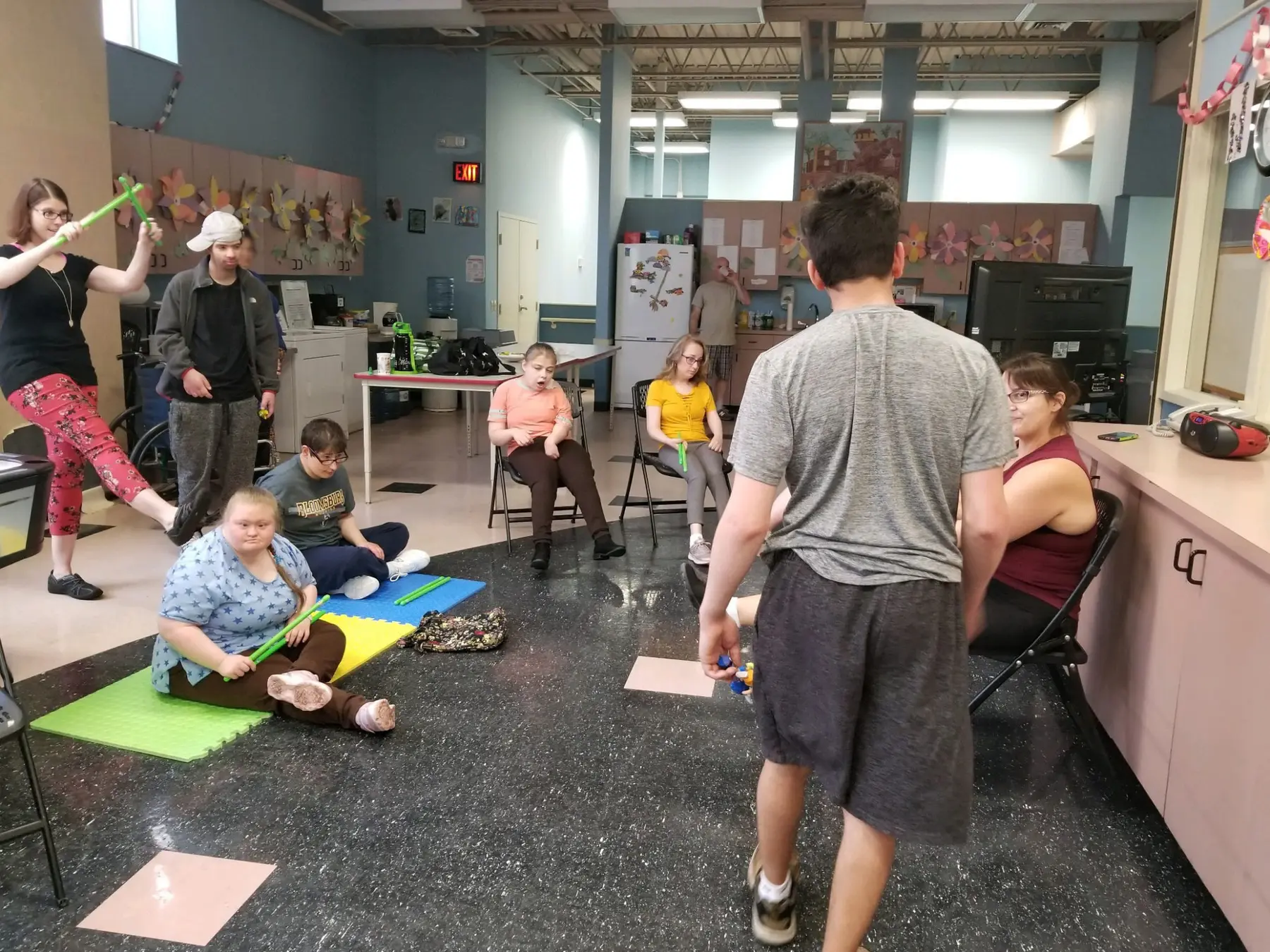 Several people sit and stand in a large room with colorful mats on the floor, holding green sticks, while one person stands in the foreground facing away from the camera.