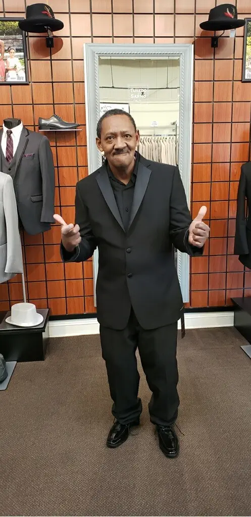 A man in a black suit stands in a clothing store giving a thumbs-up gesture with both hands.