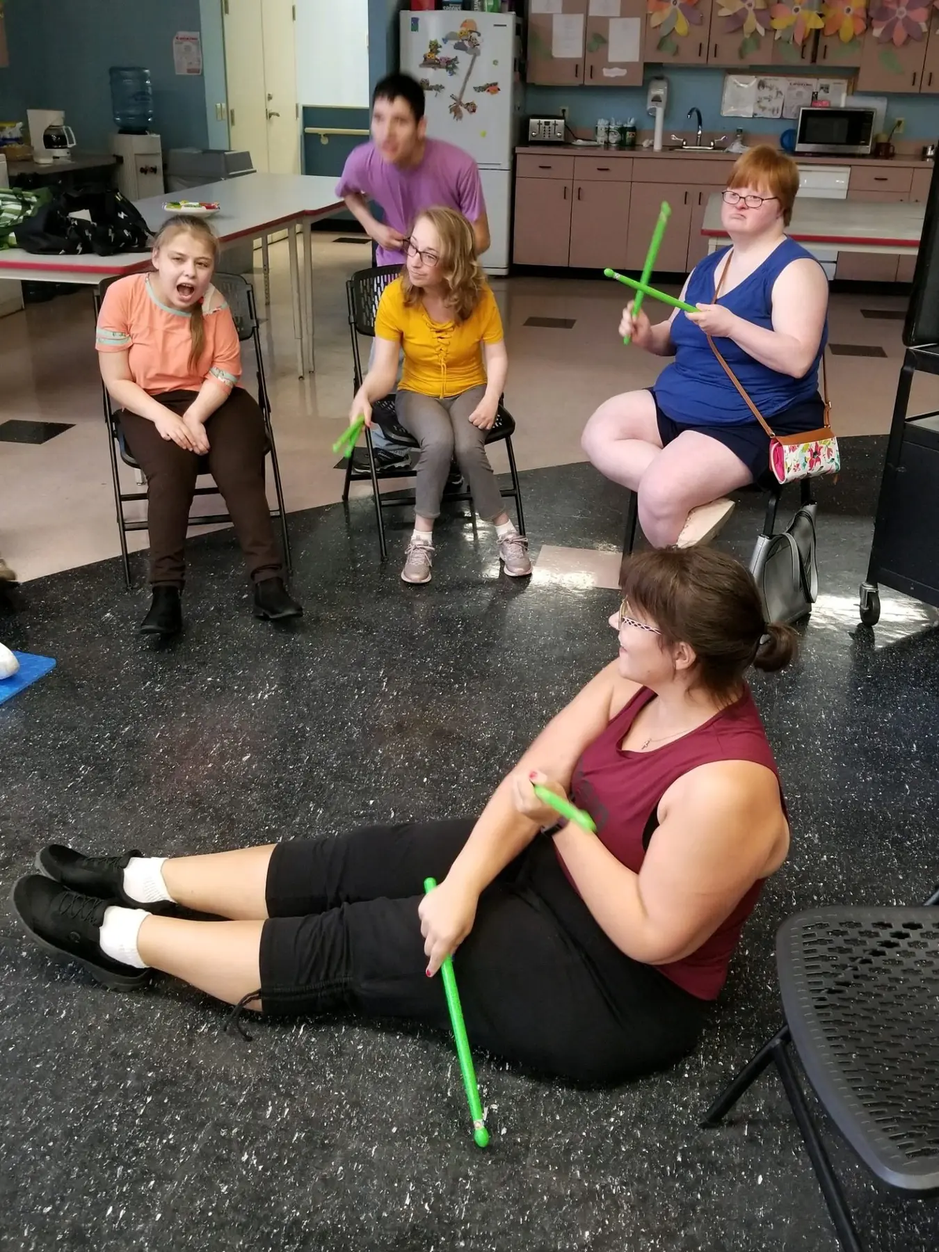 Five people sit or stand in a room holding bright green rhythm sticks, with one person seated on the floor in the foreground and others on chairs or standing behind.