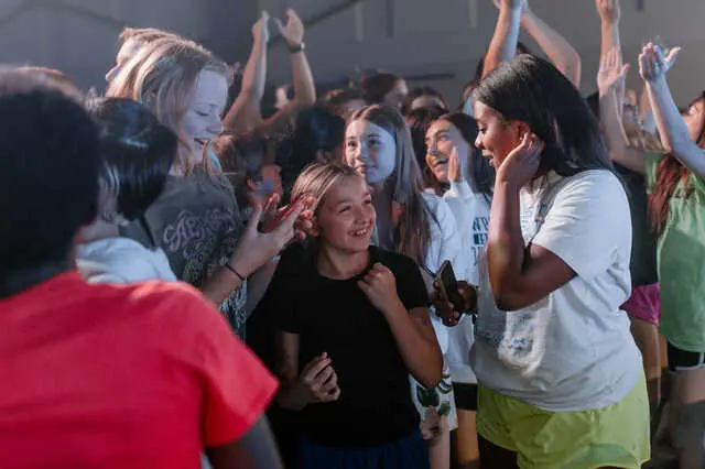 A group of children and teens stand close together indoors, smiling and raising their hands while one girl in a white shirt holds a phone near her ear.
