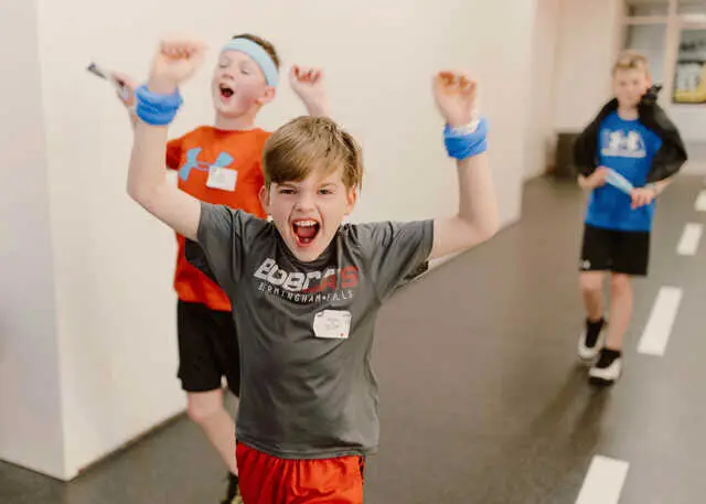 Three boys in athletic clothing stand in a hallway, with the boy in the foreground raising his arms and shouting while the others smile behind him.