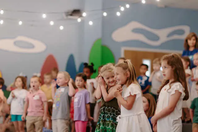 A group of children stand together in a room with painted wall shapes and string lights, with two girls in the foreground smiling and holding hands.