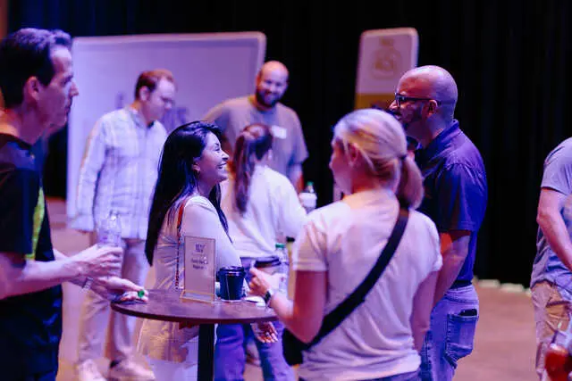 Several adults stand and talk in small groups around a tall table holding a sign and cups in a dimly lit room.