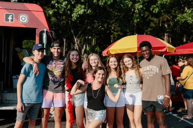 A group of young people stand together smiling in front of a red food truck with umbrellas behind them.