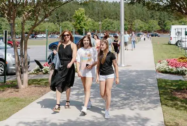 Three women walk toward the camera on a paved path lined with grass and trees, with other people and parked cars visible in the background.