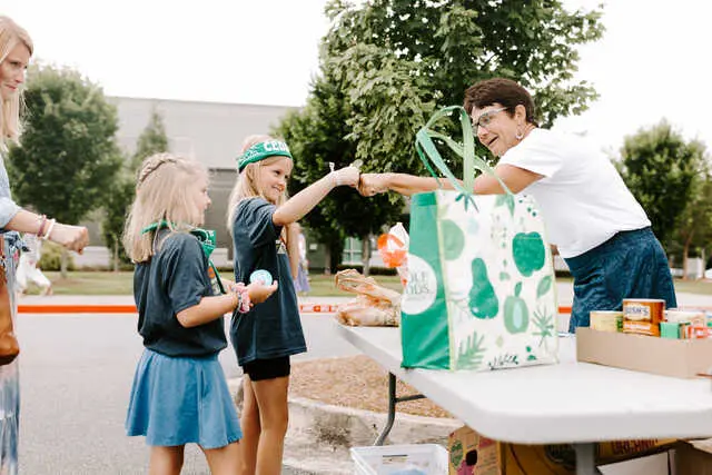 Two girls wearing green headbands and dark shirts stand outdoors and reach toward a woman holding a large green gift bag across a white table with boxed food items.