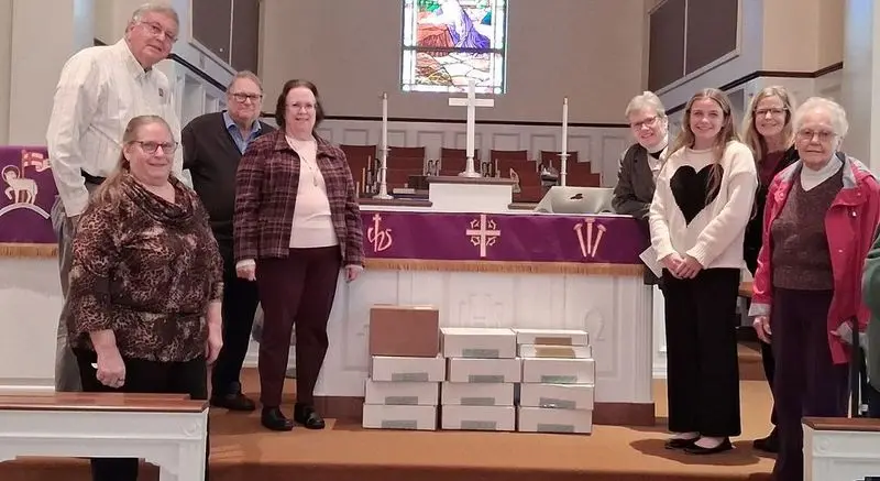 Eight people stand in a church sanctuary in front of an altar with a purple banner, cross, candles, and stacked white boxes on the floor.