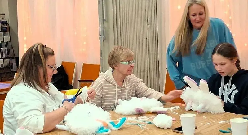 Four people sit and stand around a table assembling white fluffy bunny crafts with scissors and glue.