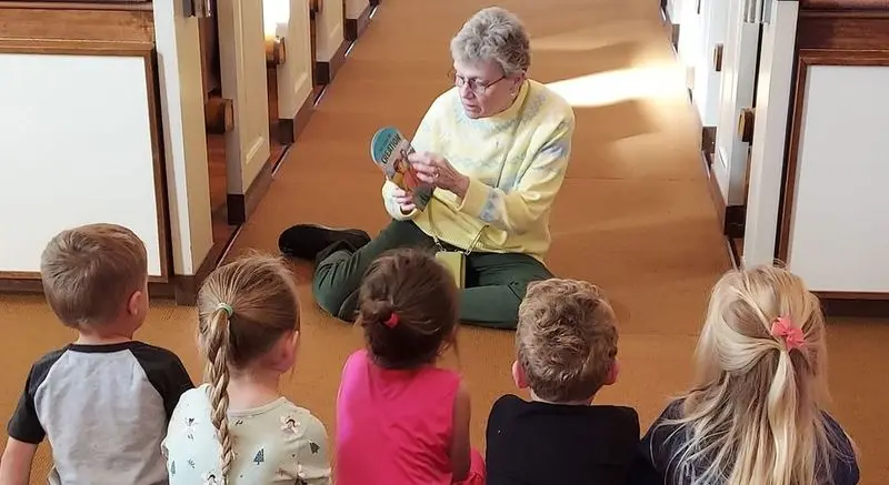 An older woman sits on a carpeted floor holding a book while five children sit facing her in a hallway.