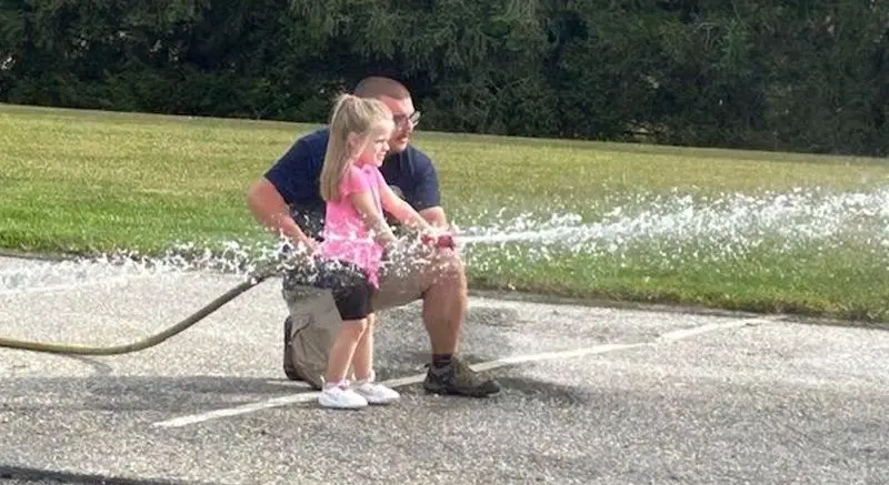 A man kneels on pavement holding a hose while a young girl in a pink shirt stands beside him and water sprays outward.