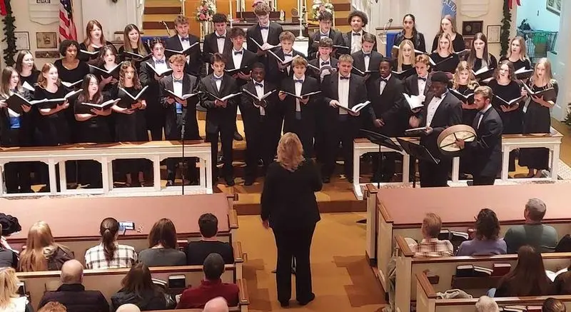 A large choir of children and adults in black robes stands on risers behind a wooden rail while a conductor directs them in a hall with an audience seated in the foreground.