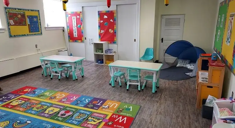 An empty childcare classroom with two small tables and chairs, a large alphabet rug on the floor, wall decorations, and a blue play tent in the corner.