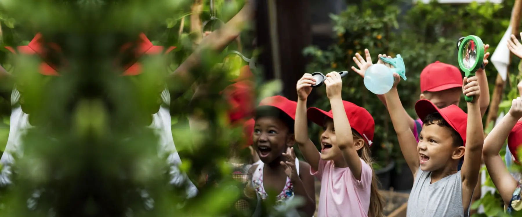 Several children wearing red caps hold magnifying glasses and a spray bottle while looking excitedly at green foliage.