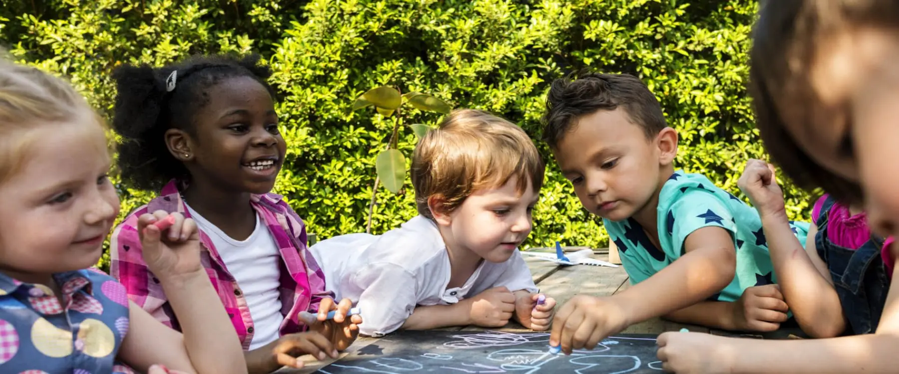 Five children sit and lean around a table outdoors, drawing with markers on a dark surface, with green bushes behind them.