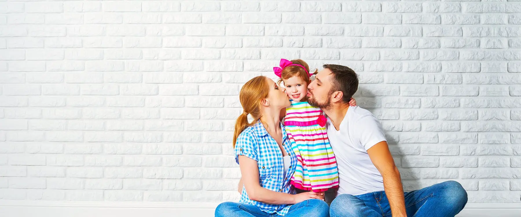 A man and woman sit on the floor against a white brick wall, both kissing the cheeks of a young girl in a striped dress and pink bow.